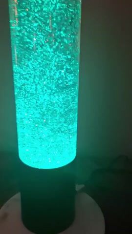A tall, clear cylindrical lamp filled with a bright turquoise liquid and shimmering particles is illuminated from the bottom. The particles swirl and dance within the liquid, creating a mesmerizing effect. The lamp sits on a dark cylindrical base, which rests on a white circular surface. The background is dimly lit and appears to be a wall. The camera is positioned in front of the lamp, capturing its full length and the dynamic movement of the particles. The lighting is soft and atmospheric, highlighting the glowing effect of the lamp.
