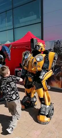An outdoor event features two individuals in elaborate robot costumes, one resembling Optimus Prime (blue, red, and silver) and another Bumblebee (yellow and black), standing on a paved surface. A young boy in a black and white patterned shirt and light-colored pants is visible on the left, observing the robots. The Optimus Prime costume makes a turning movement with its upper body. The Bumblebee costume also pivots and turns, showcasing its detailed construction from various angles. In the background, a large building with a glass facade is visible under a clear sky, along with a red tent, a white tent, speakers, and banners depicting Transformers characters. Other blurred figures and event elements are present in the distant background.