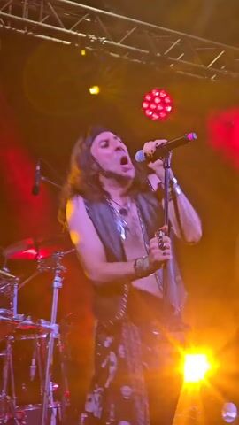 A male singer with long, dark, wavy hair and a black bandana sings into a microphone on a stage. He is wearing a black leather vest over his bare chest, black leather pants, and a black scarf with skulls tied around his waist. He has several necklaces and bracelets. A drum set is visible to the left of the singer, and stage lights are set up above. The background appears to be a stage curtain or backdrop with a snake design. The lighting is red and yellow.