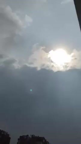 The video shows a low-angle view of the sky. Initially, the sun is visible through a layer of gray clouds, but the clouds gradually become darker and denser, obscuring the sun completely. The tops of buildings are visible at the very bottom of the frame throughout the video. There are no people visible in the video.