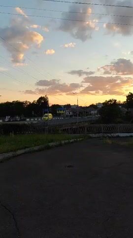 This media file captures a sunset scene in Korosten, Ukraine. The foreground features a dark, cracked asphalt surface, possibly a road or parking lot. Beyond this, a strip of green grass and a wire fence separate the foreground from a road or railway line.

Across the railway, a collection of buildings and trees are visible. A bright yellow van is driving on a road in front of the buildings. The sky is filled with dramatic clouds, illuminated by the setting sun, which casts a warm, orange glow across the horizon. The overall mood is peaceful and serene, typical of a sunset. There are several sets of power lines stretched across the sky, indicating the presence of infrastructure. The time of day is clearly late afternoon or early evening due to the sunset. The scene appears to be a somewhat rural or suburban area on the outskirts of a town.

There is a sign visible on one of the buildings in the distance, but the text is too blurry to read clearly. The image is a still photograph.