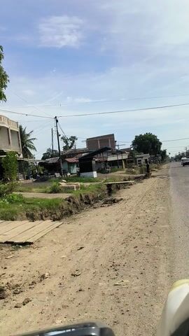 The video shows a view from a vehicle moving along a dirt road. On the left side of the road are several buildings, some appearing to be homes and others possibly small businesses. There are utility poles with wires. The ground is uneven, with some vegetation and debris. On the right side of the road, the terrain is also rough and unpaved. In the distance, more buildings and trees are visible under a partly cloudy sky. A person can be seen walking near one of the buildings on the left. The vehicle appears to have its side mirror visible on the far right of the frame.