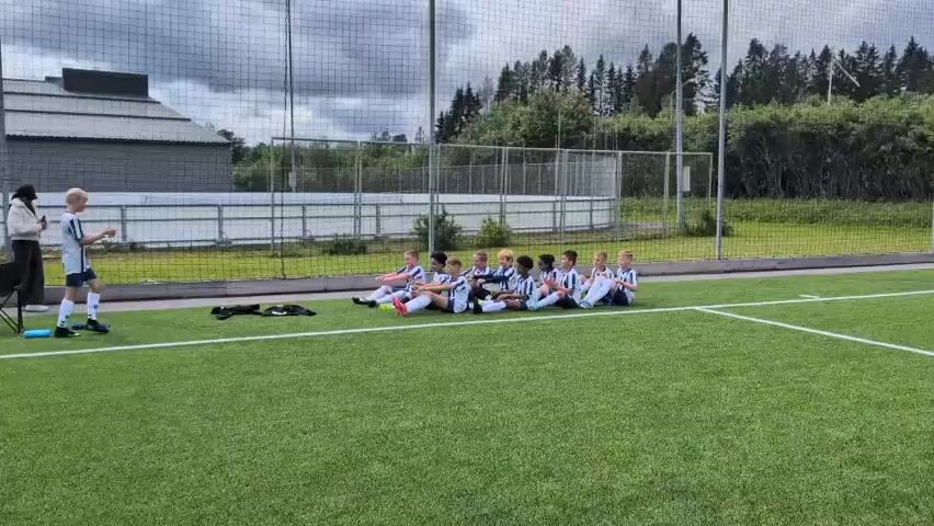 A group of young soccer players, all wearing matching black and white striped jerseys and shorts, are sitting in a line on a bright green artificial turf soccer field. They are facing to their left, with their legs extended in front of them. Their body language suggests they are attentive and possibly listening to instructions.

To the left of the seated players, a lone, blond-haired boy stands, facing the seated group. He is also in a soccer uniform, similar to the others, and appears to be addressing them or leading a drill. His posture is upright and engaged. Near him, on the grass, are some black soccer bags and a pair of black socks.

In the background, a tall chain-link fence runs along the perimeter of the field. Beyond the fence, there is a white railing, followed by a strip of grass, and then a dense line of tall green trees forming a forest. To the far left, a modern building with a dark, angular roof is visible. The sky is overcast with some patchy clouds, suggesting a possibly cool or humid day.

The overall scene depicts a soccer practice or team meeting for a youth soccer team. The environment is a typical outdoor sports facility. There are no visible text elements in the frame. The mood appears focused and disciplined, typical of a sports training session.