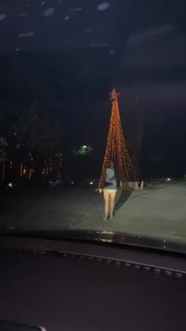 A person stands in front of a tall, cone-shaped structure adorned with numerous small, bright orange lights and topped with a large orange star. The scene is dark, suggesting it is nighttime. In the foreground, the interior of a vehicle is visible, including the dashboard, a car air freshener hanging from the rearview mirror, and a door panel with a glowing blue accent light. The person is wearing a light-colored top and light-colored shorts and appears to be looking towards the camera. Other decorative lights are visible in the background, illuminating trees and the surrounding environment. The video appears to be shot from inside the vehicle, capturing the view through the windshield.