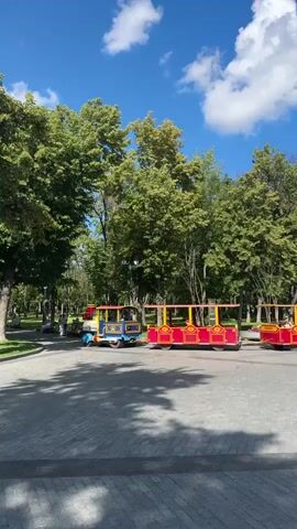 The video shows a park scene on a sunny day. There are several small, red and yellow passenger trains parked on a paved path. The trains appear to be stationary. The park is filled with lush green trees, and the sky is blue with scattered white clouds. The ground is paved with interlocking bricks, and there are shadows cast by the trees and the trains. The overall impression is a peaceful, recreational setting.