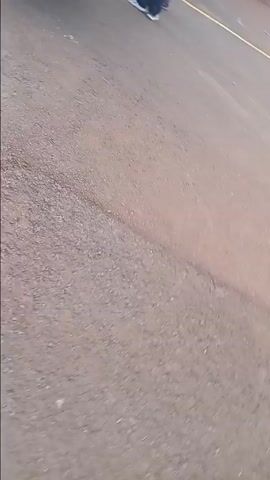 The video shows a group of people walking along a road, seemingly participating in a procession or protest. They are blowing colorful vuvuzelas and carrying a banner that says "MURANG'A COUNTY ASSEMBLY". The road is paved and marked with yellow lines. A white car is parked along the side, with speakers on the roof. A building with a yellow and green metal gate is visible in the background. The people are dressed in various outfits, including white shirts, casual wear, and some wearing hairnets. The weather appears to be sunny.