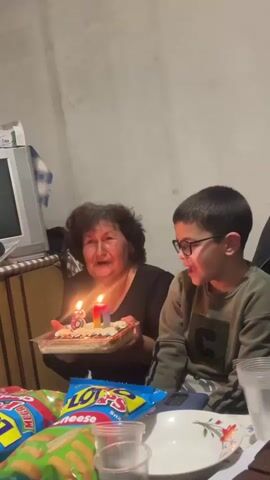 In an indoor setting, likely a home in Kosten, Bulgaria, a woman and a young boy are celebrating. The woman, with dark, curly hair and wearing a black top, holds a rectangular dessert in a clear plastic container. It has two candles shaped like the numbers "8" and "0," and another small, rainbow-colored candle, all lit. Both the woman and the boy, who wears glasses and a green long-sleeved shirt, are blowing out the candles. This activity suggests an 80th birthday celebration.

After successfully blowing out the candles, both subjects are shown laughing heartily, indicating a joyful and celebratory mood. On the table in front of them are various snacks, including two bags of chips or similar items: one blue bag clearly labeled "LOTS Cheese Snack" and another red bag with "Snacks" partially visible. There are also clear plastic cups and a white plate with a floral design. In the background, an older model silver/grey CRT television sits on a wooden cabinet, against a plain, light-colored wall. The scene captures a warm, familial moment of shared happiness.