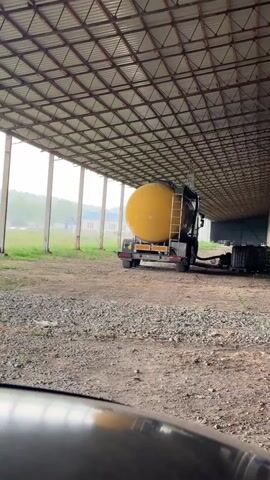A truck with a large yellow cylindrical tank is parked under a covered structure. The truck is facing away from the camera and appears to be connected to some equipment by a hose. The structure has a metal roof supported by concrete pillars. Outside the structure, there is a grassy area with trees and some buildings in the distance. A red tractor is visible in the background. The weather appears to be overcast, and there might be some rain or mist in the air. The ground inside the structure is covered with gravel and dirt. The camera is positioned low, possibly from inside another vehicle.