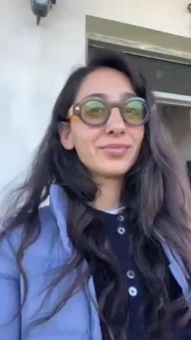 A woman stands facing the camera outside a white building. She has long, dark, wavy hair and wears round, thick-framed glasses with yellowish lenses, a navy blue cardigan, and a light blue puffy jacket. She is smiling slightly. A porch overhang and a lantern are visible above and to the sides of her. The background features a clear sky and distant landscape.