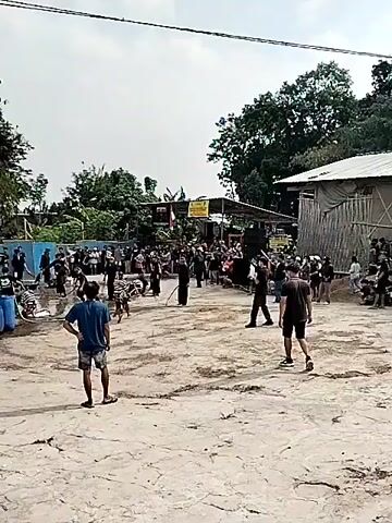 This video captures an outdoor event with a crowd gathered around a performance area. The scene is set in a sandy or dirt-covered open space, likely in a rural or semi-rural location, given the presence of trees and simple structures in the background. The sky appears bright, suggesting it is daytime, and the weather seems clear.

The central activity involves a group of performers dressed in dark clothing, some wearing traditional attire. They appear to be engaged in a performance that involves water, as there are blue barrels and hoses visible, and the ground in the performance area is wet. Some performers are on the ground, while others are standing or moving. The crowd is watching attentively, with a few individuals standing closer to the performance area.

In the background, there are buildings that look like market stalls or sheds, with some signs and flags. One sign has text that appears to be in Indonesian. There are also trees and foliage, indicating a tropical or subtropical environment. The overall atmosphere seems to be one of community gathering and cultural celebration. The people in the foreground are observers, with one person in a blue t-shirt and shorts standing with their back to the camera, watching the event.