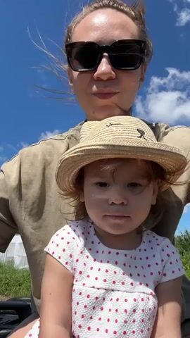 This is a close-up, first-person perspective video of a woman and a young child riding an all-terrain vehicle. The woman, wearing sunglasses and a tan t-shirt, is in the background. The child, wearing a straw hat and a white dress with red polka dots, is in the foreground, facing the camera. The child has reddish-brown hair. The sky is blue with white clouds. The overall impression is a sunny day.