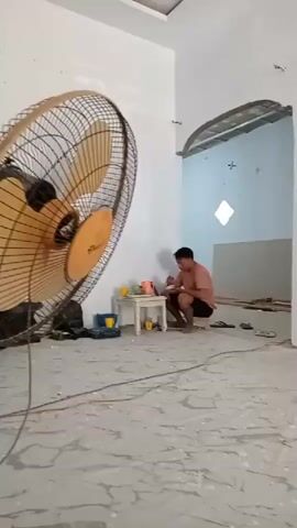 The video shows an indoor scene, possibly a house under renovation or construction. The walls are painted white. In the background, there is an arched doorway leading to another room. A wooden door with yellow trim is visible to the right of the frame. The floor is tiled with a light-colored pattern. In the left foreground, a large, circular fan is mounted on the wall. A person is sitting on a low stool or bucket in the middle ground, wearing a dark t-shirt and shorts, and a cap. They appear to be holding a phone or similar object. Another person's feet are visible in the lower right corner of the frame, suggesting the cameraman is also present. The ceiling has a modern design with recessed rectangular panels. There are some tools and containers scattered on the floor. The lighting suggests it is daytime.