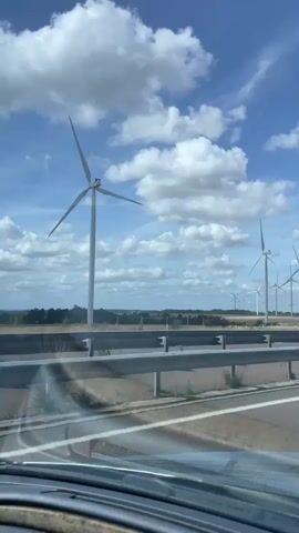 This is a video taken from inside a vehicle driving on a highway. The camera is pointed out the front windshield or side window, showing a landscape with several wind turbines in the distance. The sky is blue with scattered white clouds. The road has a metal guardrail separating the lanes. In the foreground, the dashboard and part of the steering wheel are visible. There are no people or other vehicles clearly visible in the frame. The wind turbines are large and white, with their blades rotating, indicating that they are operational. The overall scene suggests a drive through a rural or semi-rural area on a clear day.