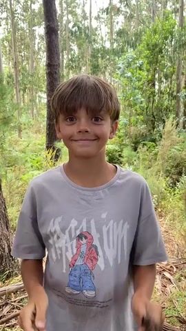 A young boy with brown hair and a grey t-shirt smiles at the camera while holding two brown seed pods on sticks. He is in a forest with tall trees and foliage. He playfully shakes his head, causing the seed pods to move.