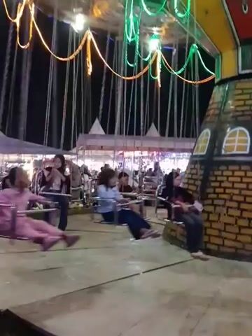 A group of people are on a carnival ride at night. The ride appears to be a swing ride with chairs suspended from a central rotating mechanism. Colorful lights hang from the top of the ride, illuminating the scene. Several people are visible in the chairs, some of whom are wearing headscarves. In the background, tents and other structures suggest a fair or festival. The ride is in motion, with the chairs swinging outwards.