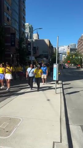 A group of people are walking down a city street on a sunny day. Most of them are wearing yellow shirts with the Colombian flag colors, likely showing support for the Colombian national soccer team. The people are walking on a sidewalk next to the street, which has cars in the background. The buildings on the left are tall, modern structures, while the buildings on the right are older, with awnings and signs. Some people are looking at their phones, while others are talking and smiling. A few people are holding drinks. The overall atmosphere appears festive and celebratory.