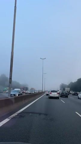 The video shows a congested highway on a foggy day. The camera appears to be positioned inside a vehicle moving slowly with the flow of traffic. The road is lined with streetlights. A variety of vehicles, including cars, vans, and a large truck, can be seen in both lanes. The fog obscures the surrounding landscape, creating a hazy atmosphere. The sky is a pale grey.