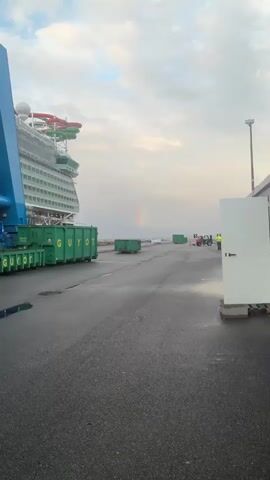 The video shows a rainbow in the sky near a cruise ship. The ship is docked at a port, and there are several green containers labeled "GUYOT" and a large blue crane nearby. A forklift and a person in a yellow vest can also be seen in the distance. The ground is wet, suggesting recent rain.
