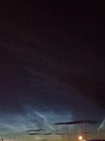 A night sky filled with dark clouds is illuminated by faint, wispy streaks of light that appear to be noctilucent clouds. The clouds are a pale blue and white, and they are arranged in delicate, ethereal patterns. The sky is otherwise dark, with only a few faint stars visible. In the foreground, the silhouettes of masts and rigging from sailboats are visible, suggesting the scene is taking place in a harbor or marina. A single bright light, possibly a streetlamp or boat light, is visible in the lower right portion of the frame.