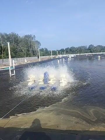 A wide shot of a large aquaculture pond, likely for shrimp farming, under a clear blue sky. In the foreground, a paddlewheel aerator churns the dark water, creating a significant splash. The pond is lined with black plastic sheeting. In the middle distance, several other paddlewheel aerators are visible in operation. Along the far edge of the pond, a fence and trees can be seen. The overall impression is one of an active and functional aquaculture operation.