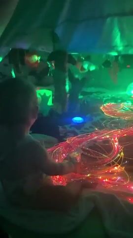 This media file showcases a dimly lit, indoor sensory play space. The primary subject is a young child, seen from behind, seated on a white blanket. The child is wearing a light-colored garment with a patterned design and is interacting with a large cluster of colorful fairy lights, primarily red and gold, that snake across the floor.

The setting appears to be a makeshift tent or fort, with a green, possibly fabric, covering overhead. The space is illuminated by a combination of the fairy lights and other ambient lighting, including a bright green light casting shadows and highlights on the ceiling and walls. The walls themselves are adorned with various objects, including what appear to be inflatable animal toys – a lion with a red face, a leopard-spotted sphere, and other animal-themed decorations. There are also hanging decorations, some of which are lit with a green glow. A patterned rug, black and white with a geometric design, covers a portion of the floor.

The overall atmosphere is one of wonder and exploration, with the vibrant lights and playful decorations creating an engaging environment for the child. The lighting suggests it is likely evening or indoors with artificial light. There is no visible text in the media file. The location context provided is Wakefield, United Kingdom, but there are no specific visual cues within the media file to confirm this.