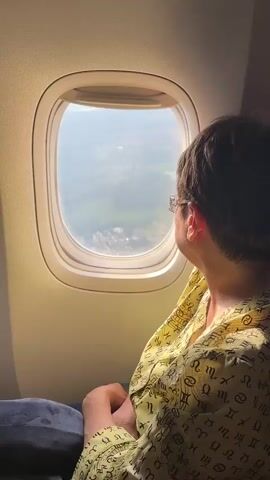 A person in a yellow shirt with a celestial print is sitting by an airplane window, looking out at the landscape below. The view outside the window shows a green, mountainous terrain with some buildings. The sun is shining, creating a bright light effect on the window. The person's right hand is visible, resting on their lap. The person is wearing glasses. The interior of the airplane is visible, including the seat cushion and the window frame. The video is shot from behind the person, focusing on their profile and the view outside the window.