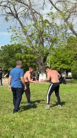 Two shirtless men in athletic pants engage in wrestling on a grassy lawn in a park. An older man in a blue shirt and dark pants stands nearby observing. Other people are sitting or standing in the background. Trees surround the area.