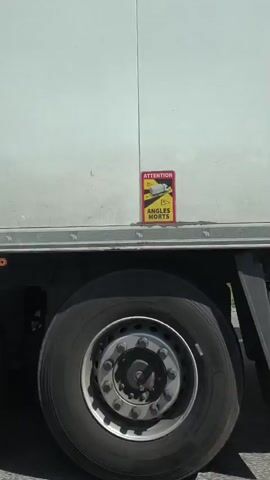 The video shows the side of a white semi-truck driving on a road. The truck has a large white cargo box and dual rear wheels. The scenery outside the truck includes trees and a blue sky with white clouds. The camera appears to be mounted on the side of the truck, capturing a motion view as it travels.