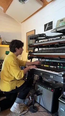 A man in a bright yellow shirt and black pants plays a stack of four keyboards in a room filled with audio equipment. He starts kneeling to play the bottom keyboard and then stands up to play a higher keyboard. There are various keyboards, amps, and other electronic devices visible, including a Fender Princeton Reverb amp. The room has white walls and a wooden ceiling with a disco ball. There are also framed prints on the wall. The man appears to be composing or practicing music.