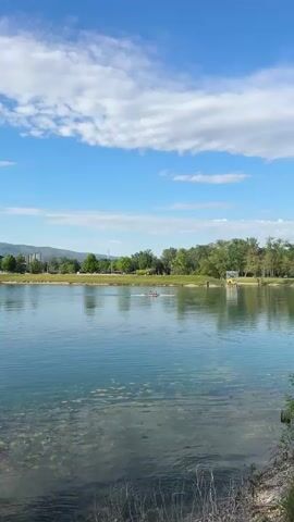 The video opens with a wide shot of a calm lake under a bright blue sky dotted with fluffy white clouds. The water is clear, revealing submerged vegetation or lily pads near the shore, and reflects the sky and distant trees. In the mid-distance, a small boat with two people rowing can be seen moving across the water.

The camera slowly pans to the left, revealing more of the lake's shoreline, which is lined with lush green trees. In the background, a range of hills or mountains stretches across the horizon, with some distant buildings visible, suggesting an urban or semi-urban area beyond the natural landscape. The overall impression is one of peace and serenity on a sunny day. The scene is consistent with a recreational lake environment, such as Lake Jarun in the city of Zagreb, Croatia, given the provided context. No text is visible in the video.