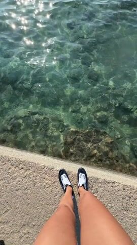 The video shows a first-person perspective of someone's legs and feet standing on a concrete or stone ledge next to clear, shallow water. The water has a rocky or sandy bottom with some darker areas that could be seaweed or rocks. Sunlight shimmers on the surface of the water, creating bright reflections. The person is wearing black and white athletic shoes. The camera angle is looking down at the feet and then pans slightly to the right, showing more of the water.