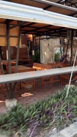 The video shows an outdoor seating area of a restaurant or cafe. There are wooden tables and benches set up on a stone patio. In the background, there is a covered structure with a corrugated metal roof supported by metal beams. Some of the seating areas are covered with what appears to be fabric or bamboo screens. There is also a white tarp covering part of the structure. A person, appearing to be a woman with light-colored hair, is seated at one of the tables in the background. In the foreground, there are several rows of purple and green plants. The overall scene suggests a casual, open-air dining environment.