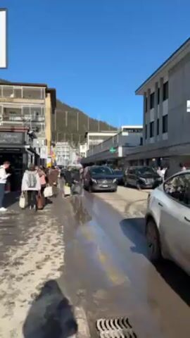 The video shows a busy street scene on a bright, sunny day in a town nestled in the mountains. Pedestrians, many wearing winter clothing, are walking on the sidewalks alongside the street. Several cars, including black Mercedes-Benz vans and a white SUV, are driving down the road. Some people are crossing the street in front of the vehicles. The road surface is wet and slushy, suggesting recent snow or melting ice. Buildings line both sides of the street, and a mountain is visible in the background. A pharmacy is identifiable by its green cross sign. The overall scene depicts a bustling urban environment in a winter resort town.
