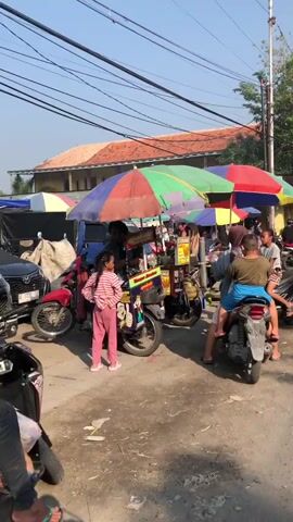 The video shows a street scene in what appears to be a market or a busy roadside area. Several food stalls with colorful umbrellas are set up along the side of the road. People are seen interacting with the vendors, purchasing food, and traveling on motorcycles. The environment is sunny with clear skies. There are utility poles and wires overhead, and buildings with tiled roofs in the background. The road surface is dirt or gravel.

In the foreground, a young girl is sitting on a black motorcycle. To her left, a woman in a pink and white striped shirt and pink pants stands near a food cart. Behind her, a vendor with a dark shirt is interacting with customers. Another motorcycle with two people on it is parked nearby.

Moving further down the street, there are more food stalls, each with its own umbrella. People are gathered around these stalls. Several motorcycles are seen driving or parked along the street. One motorcycle has two people on it, with the rider wearing a blue headscarf and the passenger wearing a pinkish-orange top. Another motorcycle is occupied by a person wearing a red hooded sweatshirt. A green motorcycle with two people on it is also visible. The overall atmosphere suggests a lively marketplace.