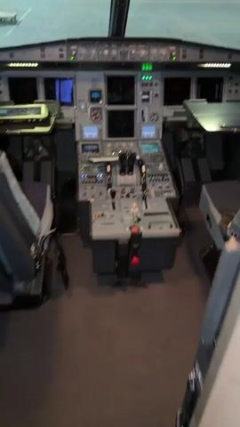 The video shows the interior of an airplane cockpit, likely an Airbus A320 series. The camera pans up from the center console, which includes the throttle controls and flight management system interface. It moves to the overhead panel filled with numerous switches, buttons, and indicators. The view continues upwards to the glare shield and then back towards the windshield, showing the view of an airport runway at night. The cockpit seats are visible on either side of the console. The lighting in the cockpit is a mix of instrument panel lights and external light from the airport. No people are visible in the video.