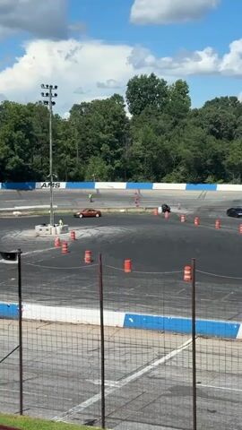 The video shows a race track with several orange traffic cones arranged on the asphalt. A brown sports car is seen drifting around the cones, with a person standing near it. In the background, a yellow bulldozer and a white pickup truck with a trailer are parked on the grass. Trees and a blue sky with clouds are visible behind the track. The scene appears to be a car event or competition.