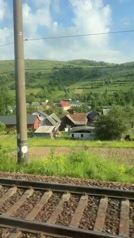 The video captures a view from a moving train. The focus is on the passing landscape, which features a small rural village nestled in a green, hilly area. Houses with varying roof colors, including red and brown, are scattered throughout the scene, surrounded by fields and trees. The sky is partly cloudy with patches of blue. In the foreground, the train tracks are visible, emphasizing the motion of the train.