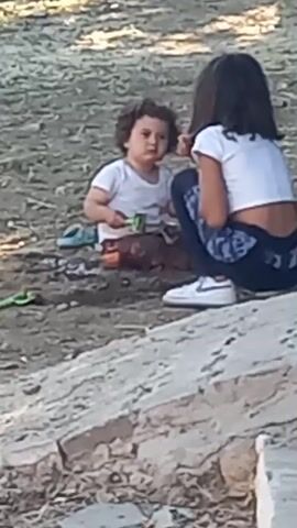 Two young children are playing on the ground outdoors. The child on the left, wearing a white t-shirt, appears to be a toddler with curly hair and a pink bow. They are holding a green object and are seated on the ground. To the right, an older child, likely a girl, is crouched down. She has dark hair and is wearing a white cropped t-shirt and dark pants with a pattern. She is looking towards the younger child. There are toys scattered on the ground around them, including a green toy to the left and some orange objects. The ground appears to be dirt or sand, and there is a concrete or stone structure in the foreground. The lighting suggests it is daytime.