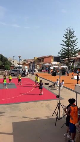 The video shows an outdoor event likely a basketball tournament. In the foreground, a group of young people are visible, some wearing orange shirts. Behind them, a bright red basketball court is set up with a few individuals engaged in play. Further in the background, an orange court is visible, with more people participating and spectating. Tents and structures suggest a festive or organized gathering. The environment appears to be a town with buildings and a large tree. The sky is clear and the lighting suggests daytime.
