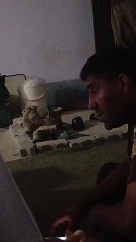 The video shows an interior scene with a person seated in the foreground, partially obscuring the view. Behind them, there's a low brick-like structure, possibly a makeshift hearth or platform, on which various objects are placed. These include a pair of dark boots stacked on a container, a white bucket, and several dark-colored bags. The walls of the room appear to be a light, neutral color. The lighting in the video is subdued. There's background noise of people speaking.