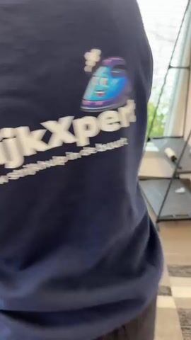 The video shows a close-up of a person's back wearing a navy blue sweatshirt with white lettering that says "Strijk". The scene then shifts to a wider shot of the same person ironing a white shirt on a blue ironing board. Several bottles of laundry products are visible on the floor next to the ironing board. The person is holding the iron and moving it across the fabric of the shirt. The environment appears to be indoors, possibly a laundry room or a home.