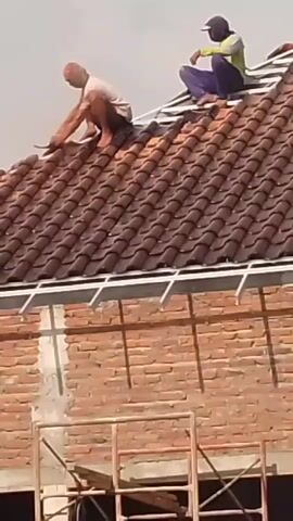 Two individuals are working on a roof. One person, wearing a light-colored shirt and orange head covering, is crouched on the left side of the roof, appearing to use a tool on the roofing material. The other person, wearing a lime green shirt and dark pants, is crouched on the right side of the roof, and appears to be arranging or installing roofing tiles. The roof is covered in reddish-brown tiles and has exposed metal framing underneath. Below the roof, a partially constructed brick wall is visible, along with scaffolding. The sky is overcast.