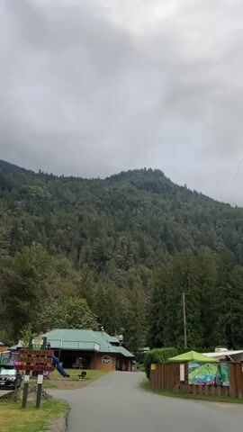 The video pans from right to left, showing a wooded mountainous landscape under a cloudy sky. In the foreground, there is a paved road with a few parked cars. To the left of the road is a building with a green roof, possibly a store or restaurant, and a playground with a blue slide. To the right of the road, there is a wooden fence and some signs. The overall scene appears to be a rural or recreational area.