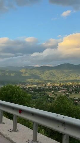 The video presents a panoramic view of a valley and a village, taken from a slightly elevated perspective, likely from a moving vehicle as suggested by the sound and the guardrail in the foreground.

In the immediate foreground, a grey metal guardrail is visible, with lush green trees and foliage below it. The camera pans from left to right, revealing an expansive rural landscape.

The midground features a village with numerous houses scattered across green fields and surrounded by trees. Many houses have red-tiled roofs, while others have lighter-colored roofs. Agricultural plots or fields are visible between the houses. In one section, several cars are parked near a building. Further into the pan, a prominent large building with a bright red roof stands out in the midground, possibly a barn or a larger facility.

The background is dominated by rolling green mountains, stretching across the horizon. The mountains are covered in trees and show varying shades of green, with some areas appearing darker due to shadows and others illuminated by the sunlight. In the distant right, more rugged mountain peaks can be seen.

The sky is partially cloudy, with a mix of bright blue patches and large, fluffy clouds. Some clouds are illuminated with golden light, suggesting late afternoon or early evening, while others are grey. The lighting creates a contrast of light and shadow across the landscape.

The overall scene depicts a tranquil, picturesque village nestled within a mountainous region, characteristic of Miločani, Montenegro. No visible text is present in the video.