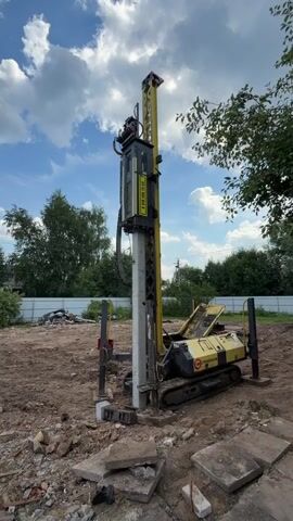 This video shows a construction site where a pile driver is being operated to install a concrete pile. A shirtless man, wearing shorts and a cap, is seen on the left side of the frame, interacting with the machinery. The pile driver, a yellow and black tracked vehicle, is positioned next to a tall concrete pile that is being driven into the ground. The machine has a large yellow mast and a black power unit at the top. The surrounding area is a dirt lot with debris, including broken concrete slabs, scattered around. In the background, there is a white fence, trees, and houses, suggesting a semi-rural or suburban setting. The sky is partly cloudy with a blue sky visible, indicating daytime. The overall atmosphere suggests active construction work.

Visible text on the pile driver includes a yellow label with black text that appears to be a serial number or model information, "B 20 466 01-1". There is also a red and white logo with Cyrillic text that reads "СТРОЙМАШИНА" (StroyMashina), which likely identifies the manufacturer or brand of the equipment.