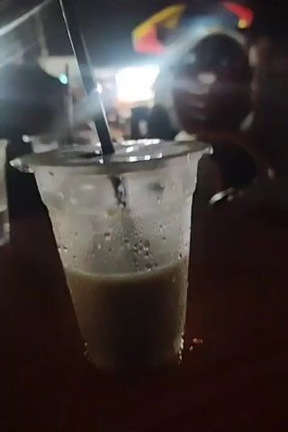 A close-up video shows a plastic cup filled with a light brown liquid, likely a beverage, sitting on a dark surface, possibly a table. Condensation covers the outside of the cup. A black straw is visible inside the cup. In the background, there is another clear plastic cup with liquid, and blurry lights and shapes suggest an outdoor or semi-outdoor setting at night. There is no discernible human interaction in the frame.