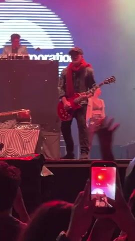 A live musical performance is taking place on a stage. In the foreground, a crowd of people is visible, with one person holding up a smartphone to record the event. On the stage, a male guitarist wearing a cap, scarf, and vest is playing a red electric guitar. To his left, another person is seen behind a console, possibly a DJ or keyboardist. A female performer in shimmering pants and a black top stands to the right of the guitarist. The stage is lit with vibrant red and green lights, and a large screen behind the performers displays a graphic with the word "poration".