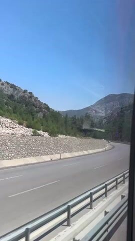 A view from a moving vehicle shows a paved road winding through a mountainous landscape under a clear blue sky. On the left side of the road, a stone retaining wall supports a hillside covered with trees and rocks. Several vehicles, including trucks and cars, are visible on the road. The camera perspective is from inside the vehicle, looking out the side window.