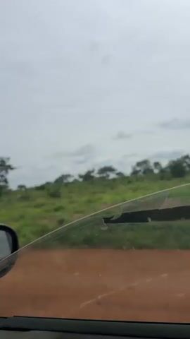The video shows a view from inside a car, looking out the passenger window. The car is moving on a dirt road. The landscape outside is rural, with rolling green hills covered in vegetation and scattered trees. The sky is overcast with clouds. The window has a wiper on it, and there are some scratches or marks on the glass. The interior of the car is visible at the bottom and left of the frame. The overall impression is of a journey through a natural, undeveloped area.
