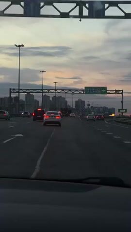 The video shows a view from inside a vehicle traveling on a highway during twilight. The sky displays a mix of gray clouds and hues of pink and orange, indicating sunset or sunrise. Several cars are visible on the highway, moving in the same direction as the camera's vehicle. On the left side of the highway, there are tall power line towers. In the distance, a city skyline with buildings can be seen. Traffic signs are present above the highway, with one sign showing directional arrows and text in a foreign language, likely indicating directions to a city or route. The interior of the vehicle is partially visible in the foreground, including a dashcam mounted on the windshield. The overall scene depicts urban traffic under a dramatic sky.